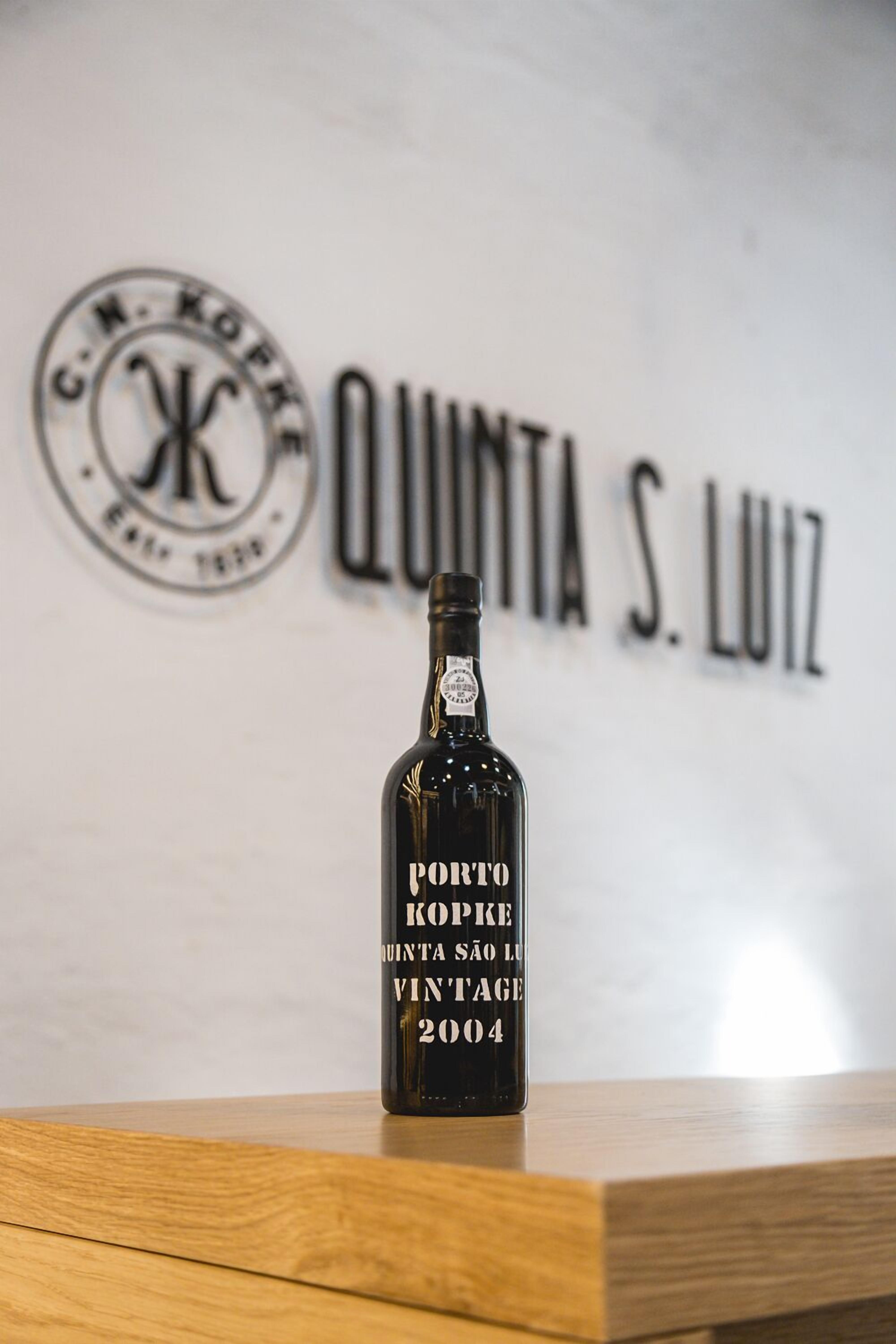 A bottle of Porto Kopke vintage wine from 2004 on a wooden surface with a backdrop featuring the text "QUINTA S. LUIZ" and a logo.