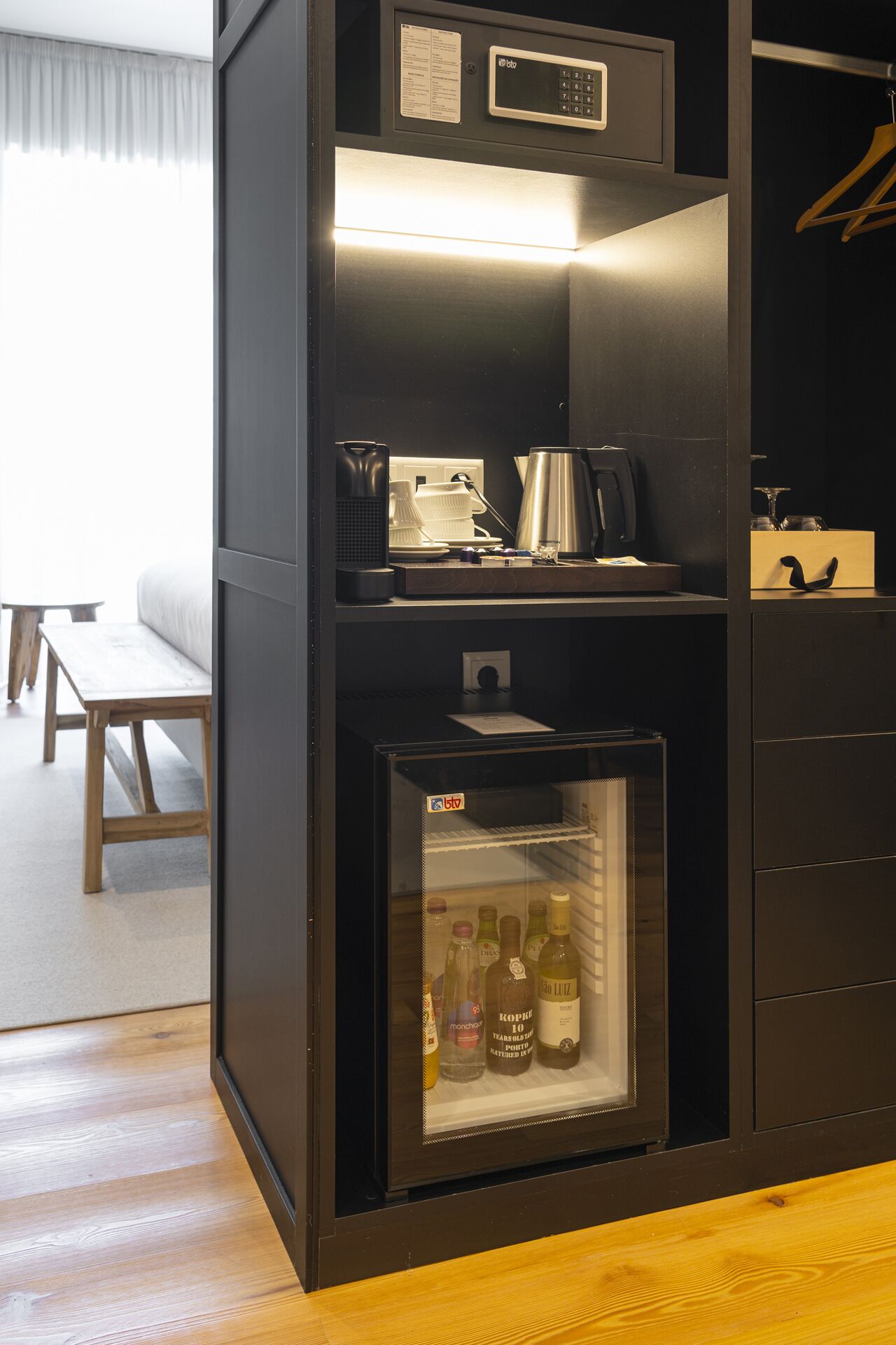Quarto compacto com minibar embutido, pequena geladeira e área de estar com mesa e banco.