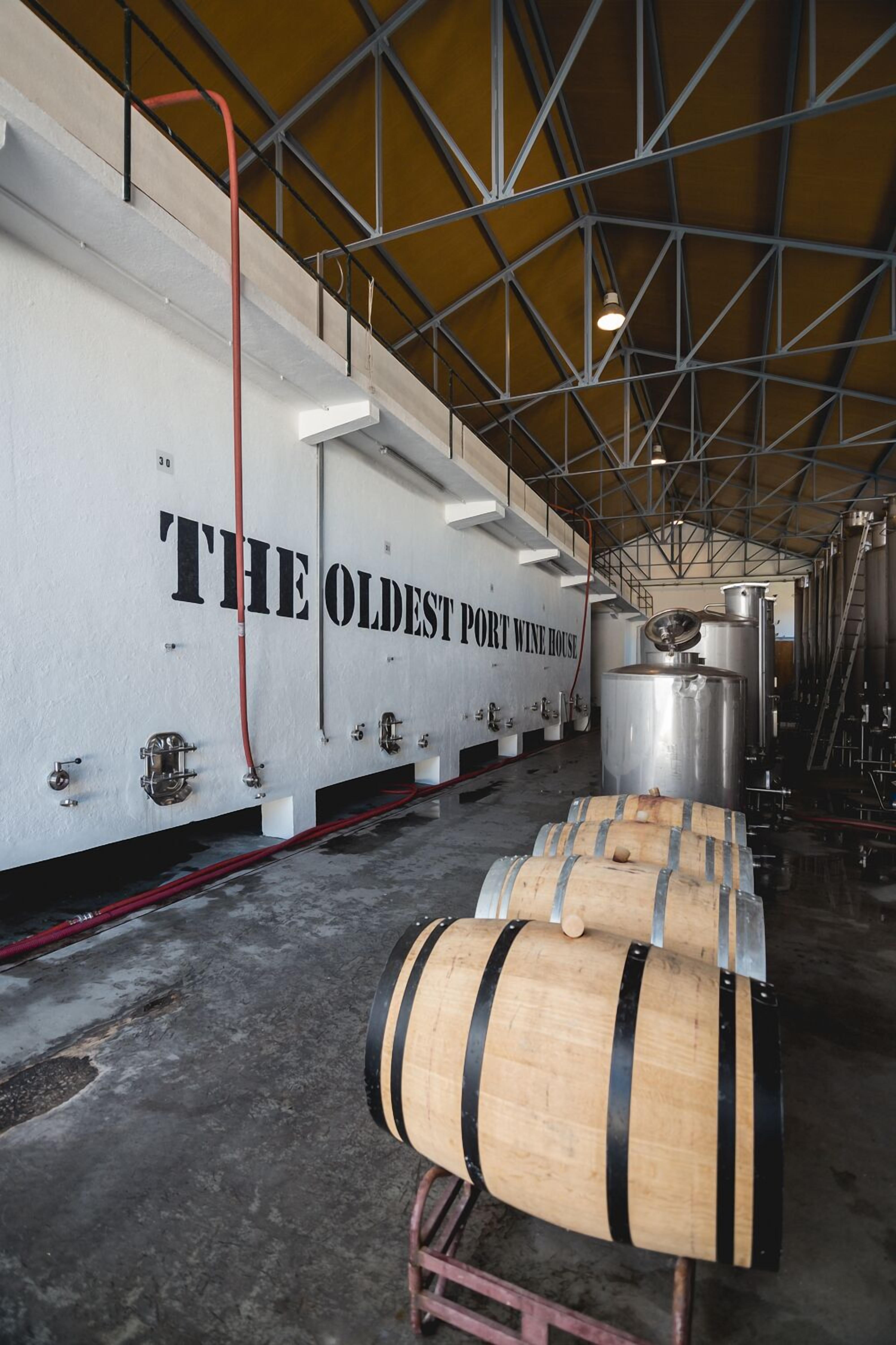 Un vaste espace industriel abritant de grandes cuves en acier inoxydable et des barriques en bois, portant l’inscription « LA PLUS ANCIENNE MAISON DE VIN DE PORTO ».