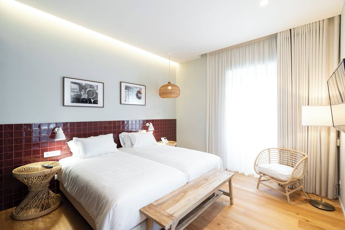 Ein Hotelzimmer mit zwei Betten, einer hölzernen Bank, einem Korbsessel und einem großen Fenster mit Vorhängen.