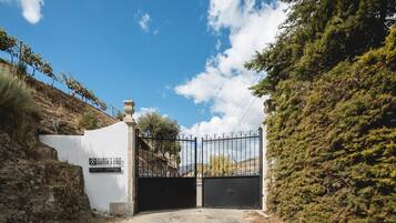 A gated entrance with a white wall and a metal gate, surrounded by trees and a clear sky.