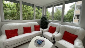 Un salon moderne avec des canapés en cuir blanc et des coussins rouges, une table basse avec des télécommandes, et de grandes fenêtres offrant une vue sur la verdure et les bâtiments environnants.
