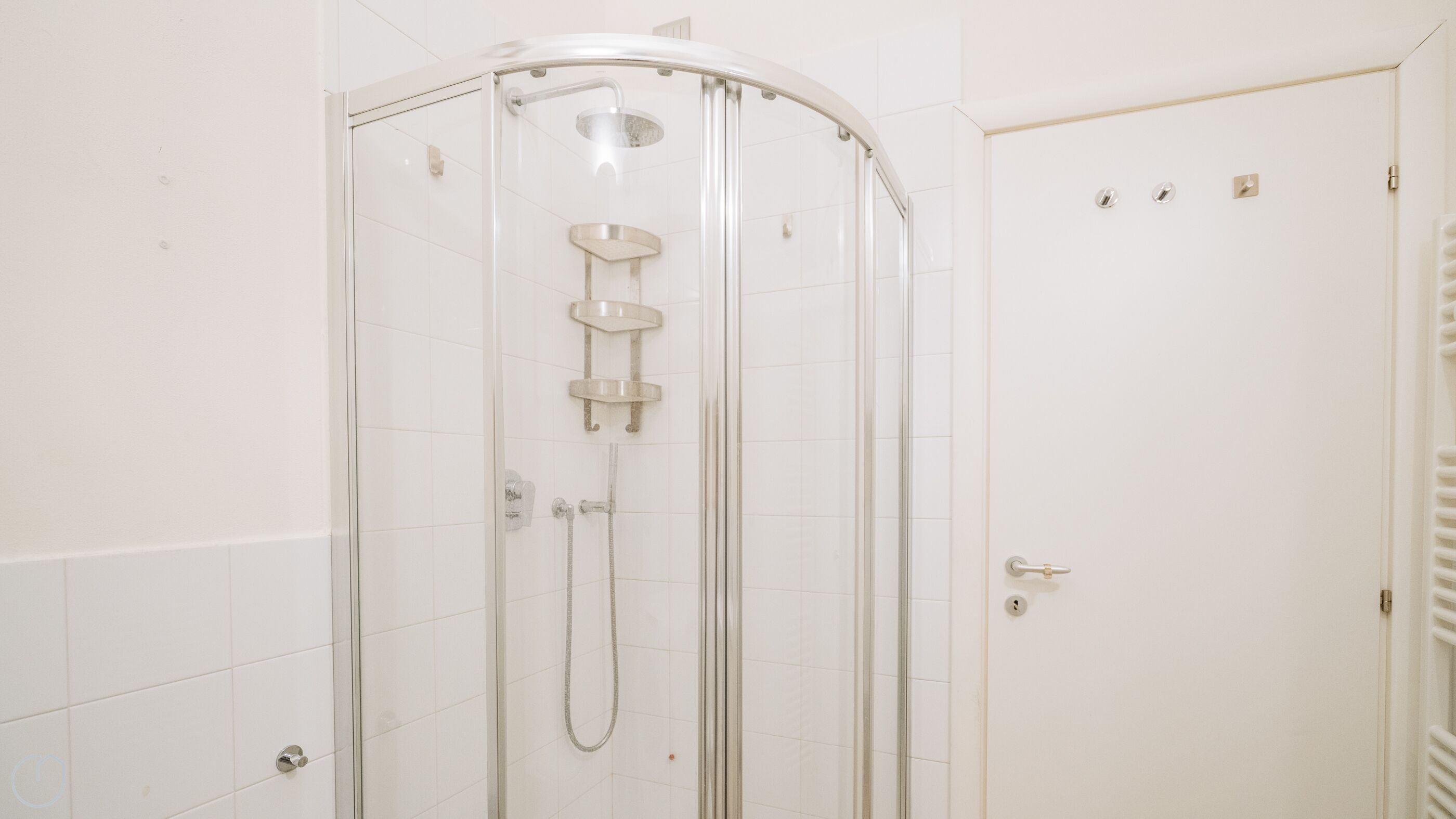 A glass-enclosed shower with a curved door, white tiled walls, and a door with a handle and lock.