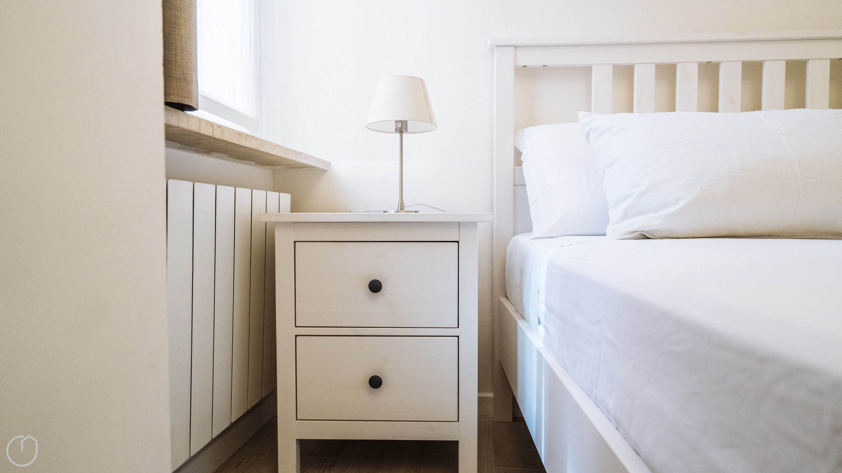 A bedroom with a white bed, a bedside table with a lamp, and a radiator.
