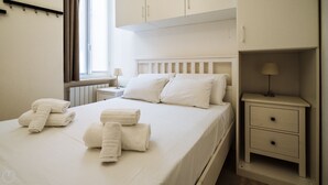 Un lit bien fait, avec du linge de lit blanc et des oreillers, une table de chevet avec une lampe et une petite commode.
