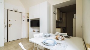 Un petit coin repas avec une table blanche dressée pour deux personnes, un téléviseur fixé au mur juste au-dessus, et une cuisine visible à travers une porte ouverte.