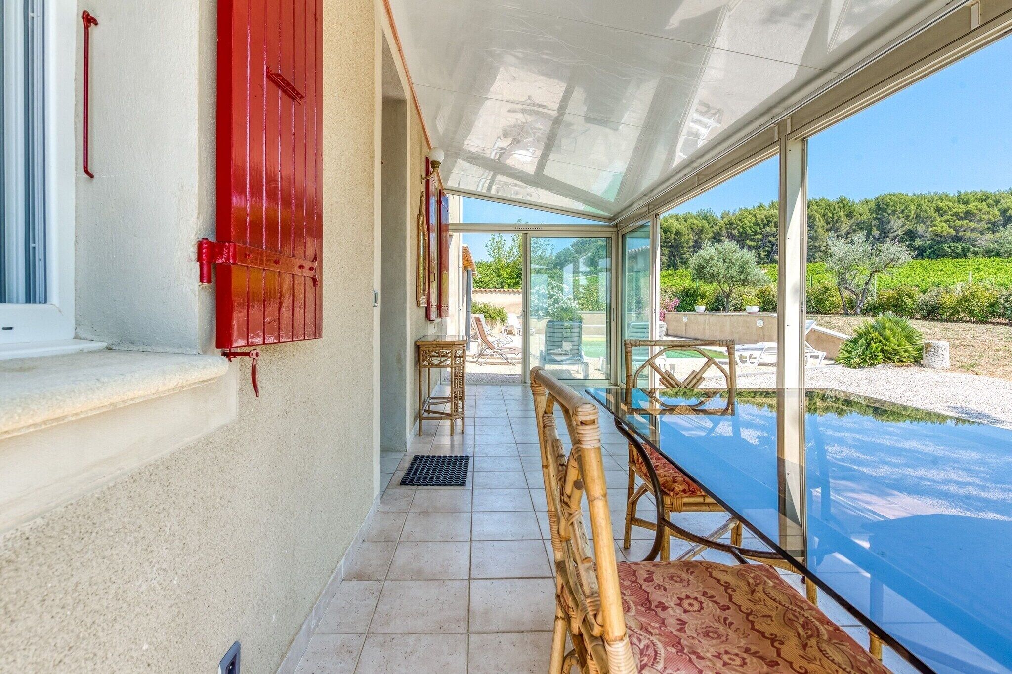 Une pièce baignée de lumière, avec une table et des chaises en verre, une porte rouge et une vue sur un jardin et des collines.