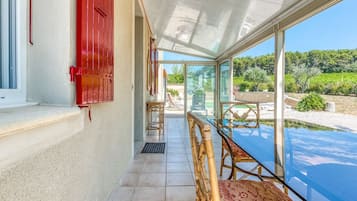 A sunlit room with a glass table and chairs, a red door, and a view of a garden and hills.