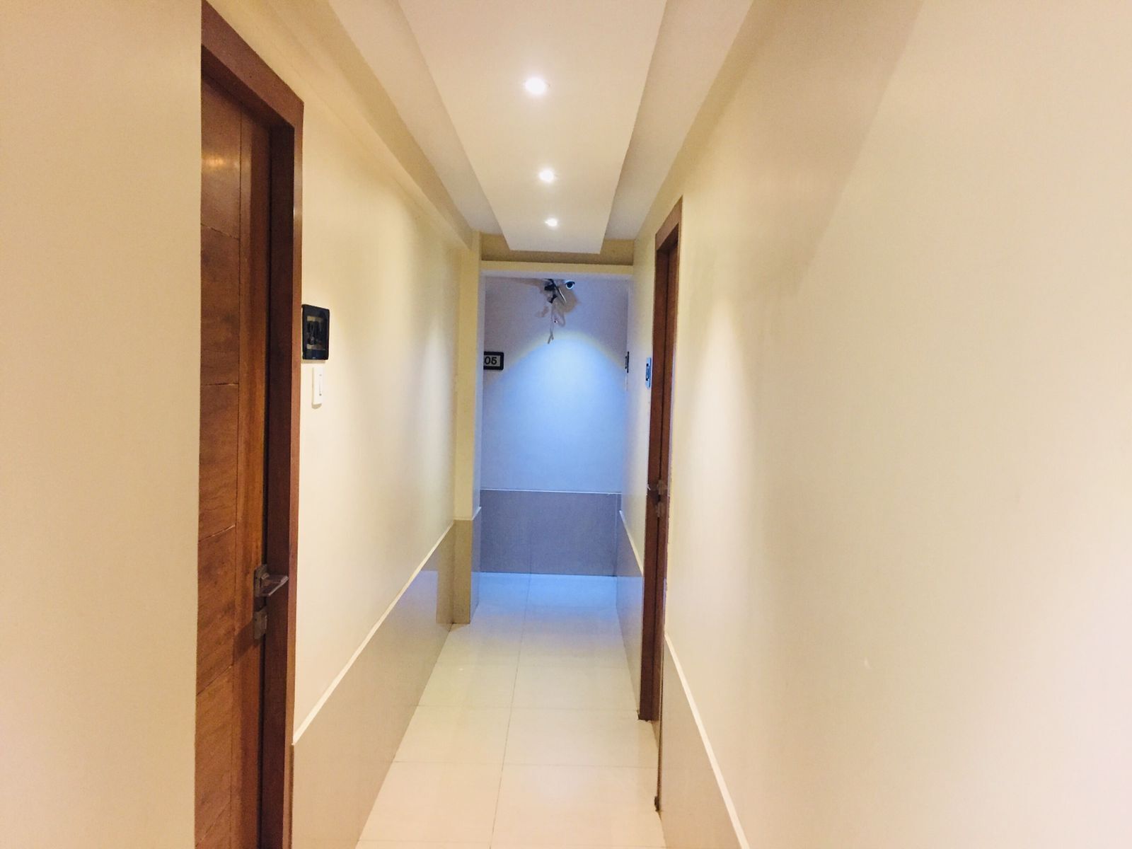 A narrow hallway with light-colored walls and a tiled floor. There are two wooden doors on either side, one of which has a sign above it. The hallway leads to a room with a ceiling fan and a light fixture.