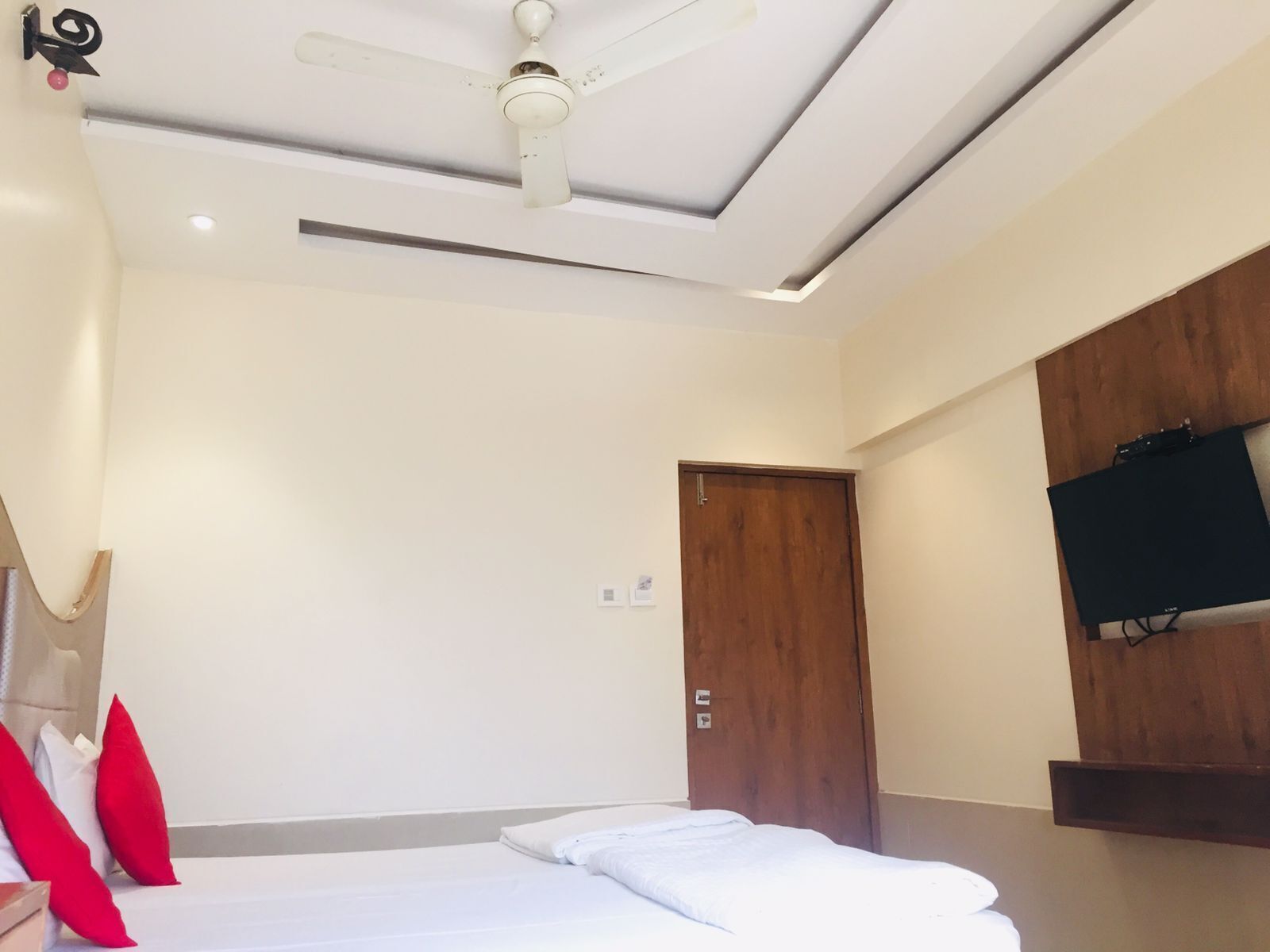 Ein Hotelzimmer mit einem Bett, einem Deckenventilator, einer Holztür und einem an der Wand befestigten Fernseher.