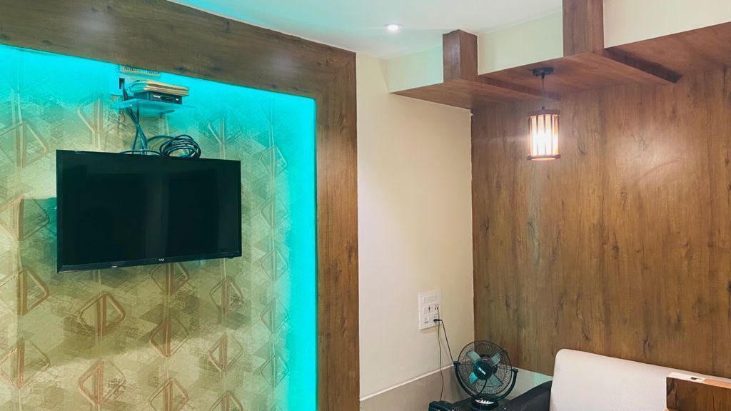 A room with a flat-screen TV mounted on a wall with a geometric pattern, a wooden ceiling, and a white wall with a power outlet and a fan.