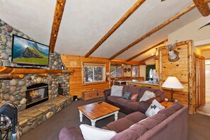 A rustic interior with a stone fireplace, wooden beams, a television mounted on the wall, and a deer head decoration.