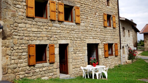 Ein Steinhaus mit hölzernen Fensterläden, einem weißen Tisch und Stühlen davor sowie einem kleinen Garten.
