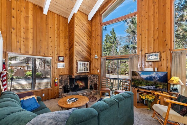 A cozy cabin interior with a stone fireplace, wooden walls, and large windows offering a view of trees.
