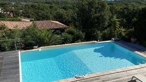 A rectangular swimming pool with clear blue water, surrounded by a wooden deck. In the background, there are lush green trees and a view of distant hills. - Villa between sea and mountains 15 minutes from the beach (zonza)