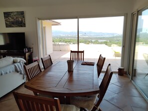 Ein Essbereich mit Holztisch und Stühlen, einer Couch, einem Fernseher und Blick auf eine gebirgige Landschaft durch große Schiebetüren.