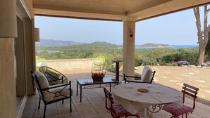 A covered patio with a table and chairs, a wicker chair with a cushion, and a view of the landscape. - Villa between sea and mountains 15 minutes from the beach (zonza)
