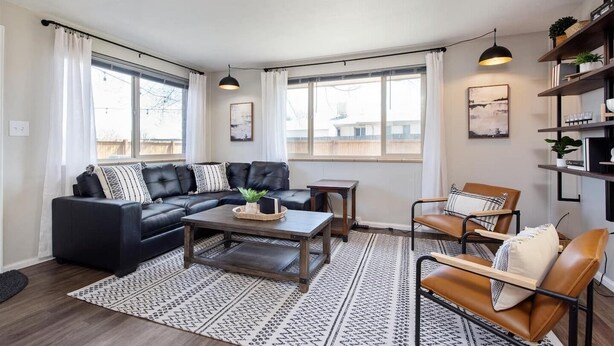 A modern living room with a sectional sofa, a coffee table, a side table, and a chair. There are two windows with curtains, a ceiling light, and a bookshelf with plants. - Modern Chic Retreat | Near Mtns & Red Rocks | Wifi (Wheat Ridge)