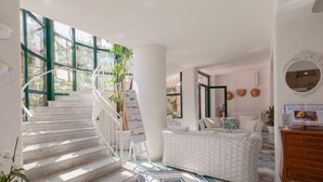 Una habitación amplia con una escalera blanca que lleva a un balcón, una cama blanca con cabecera estampada y un aparador de madera.