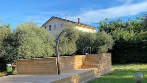 Une terrasse en bois avec des marches menant à une maison, le tout entouré de verdure et sous un ciel dégagé.