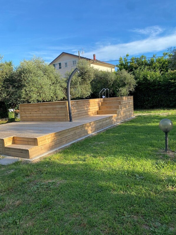 Une terrasse en bois avec des marches menant à une maison, le tout entouré de verdure et sous un ciel dégagé.