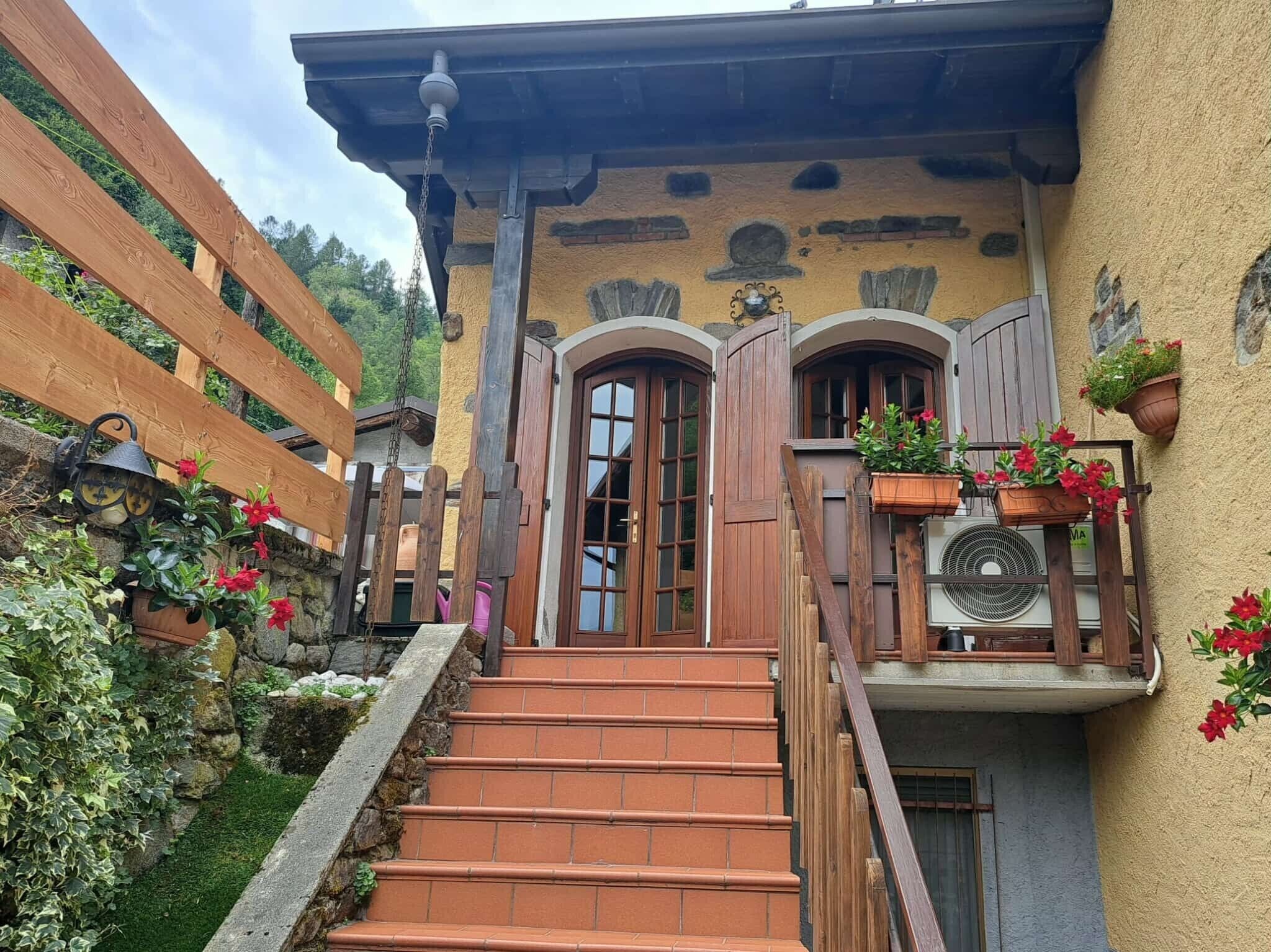 A traditional chalet with a stone facade, wooden steps leading to a wooden door, and potted plants on the balcony.