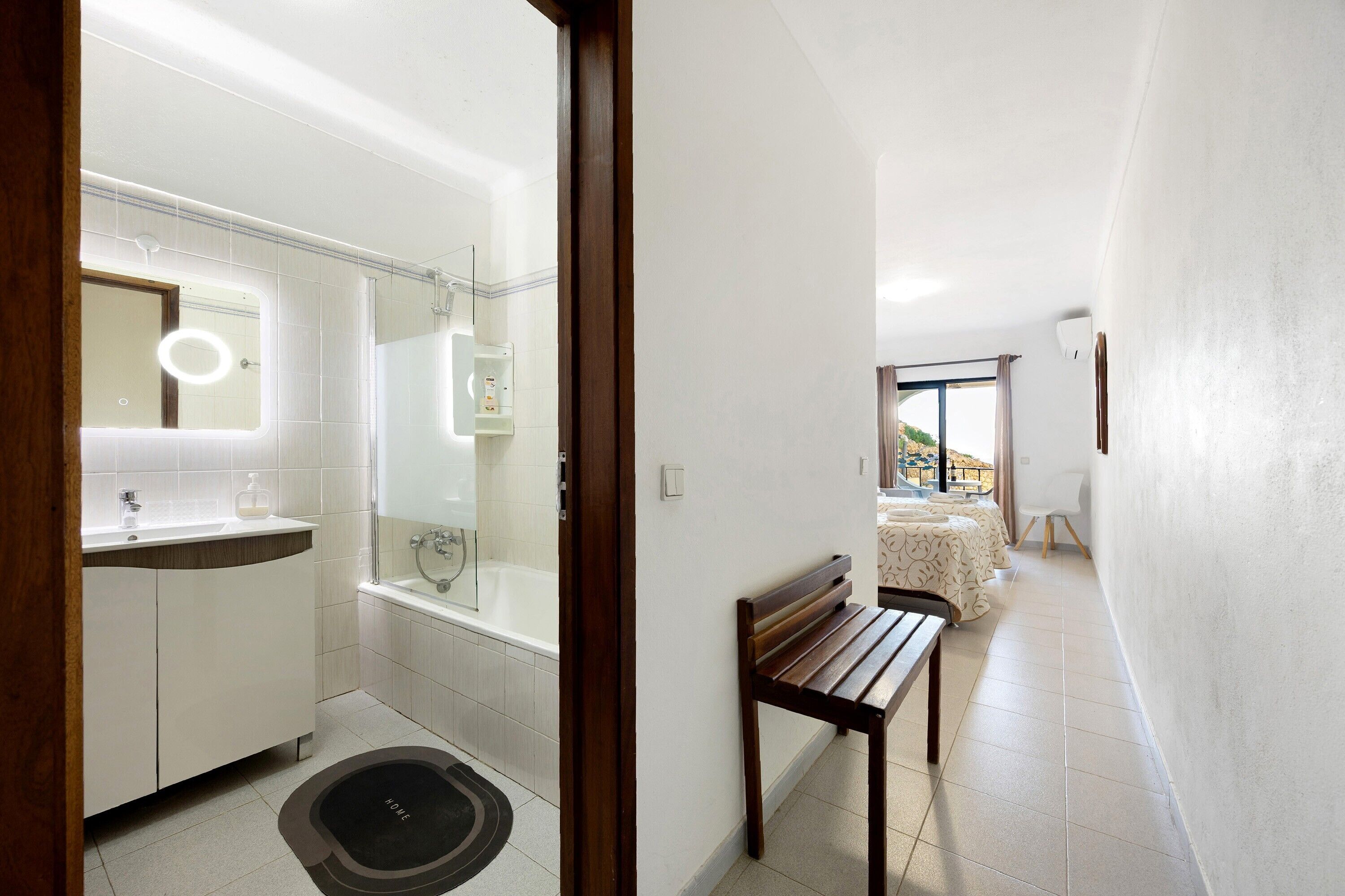A modern bathroom with a bathtub, a vanity with a mirror, and a bench in a hallway.