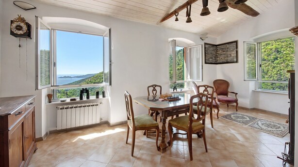 A dining area with a table, chairs, a view through a window, a clock, and a stone wall.