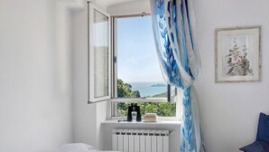 A bedroom with a bed, a window with a view of greenery and the sea, a radiator, and a chair.