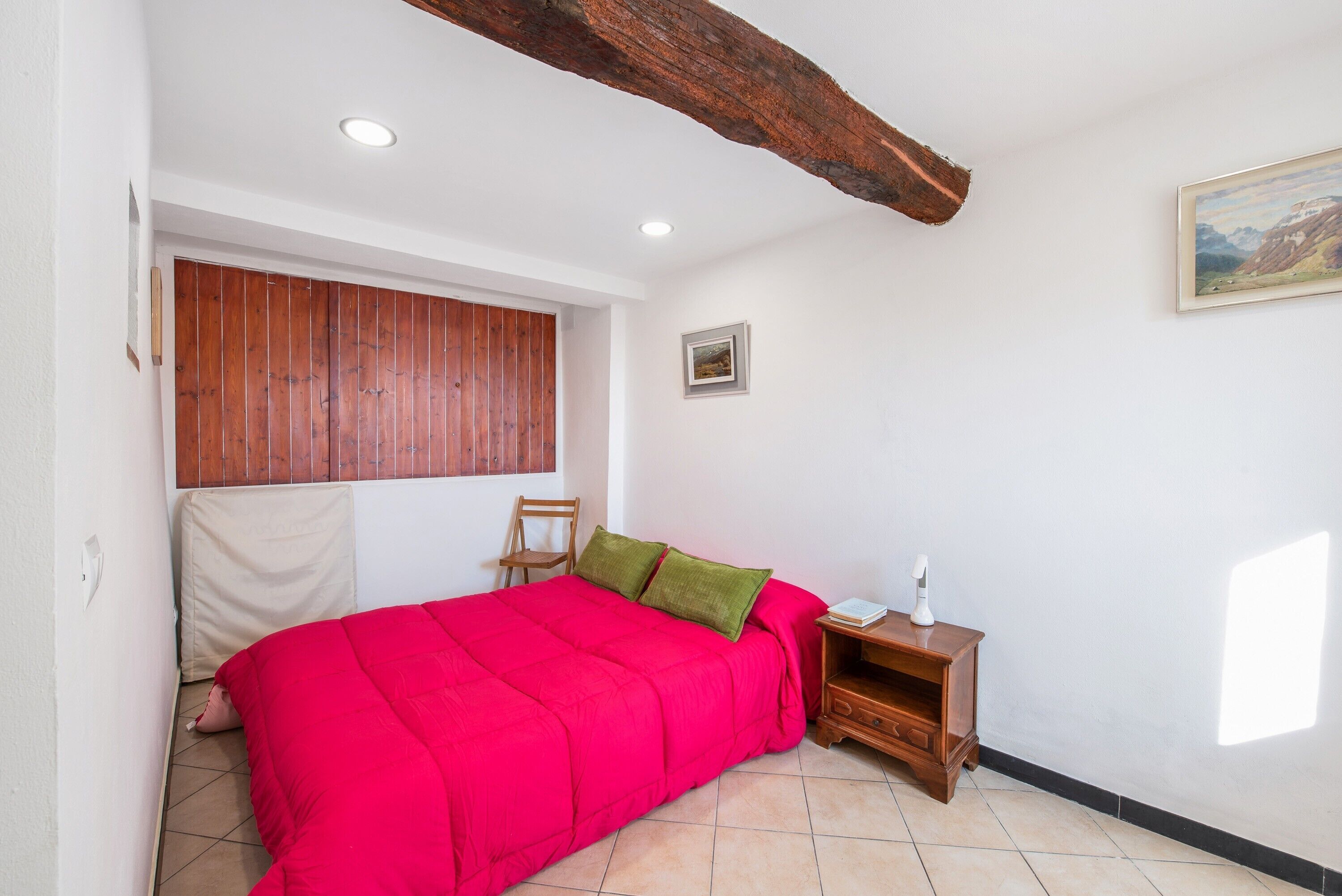 A bedroom with a red bed, wooden headboard, a nightstand, a chair, and a painting on the wall.