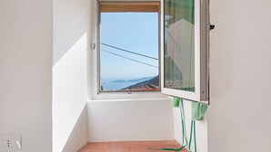 Um quarto pequeno com janela, cortina verde, piso ladrilhado e uma tomada visível.