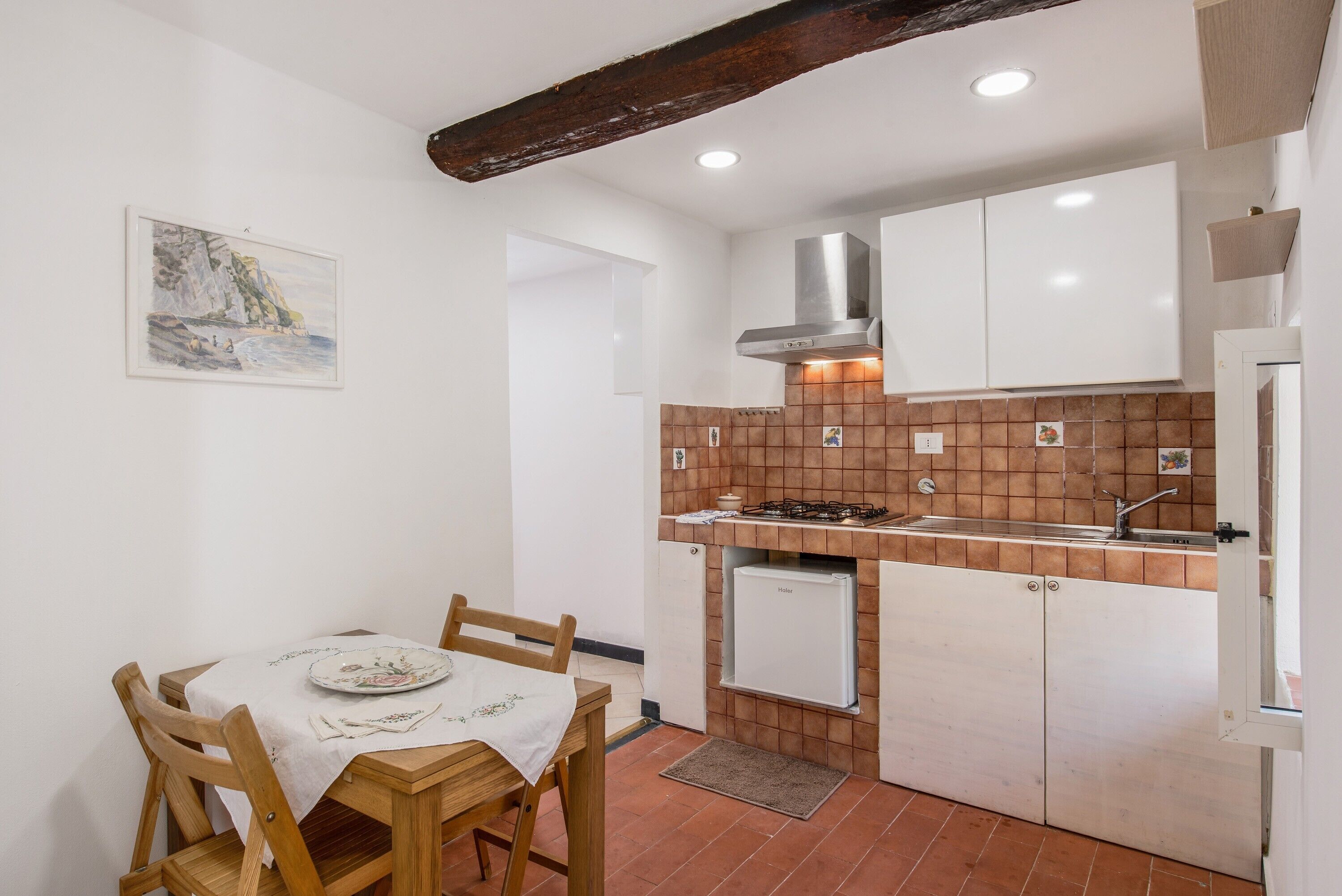 Una cucina compatta con mobili bianchi, zoccolo in piastrelle, piano cottura a gas e piccolo frigorifero.