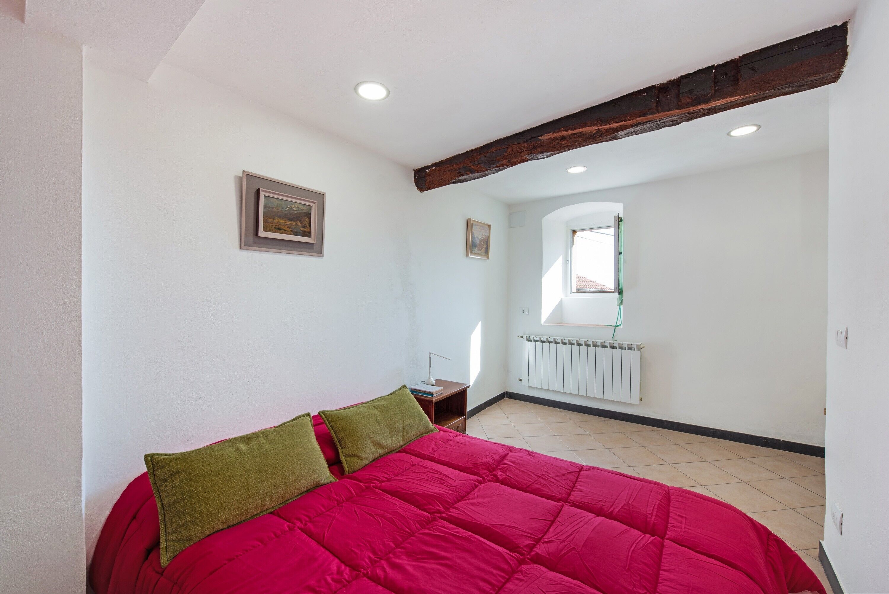Une chambre avec un lit rouge, un plafond avec une poutre en bois, un radiateur et une fenêtre.