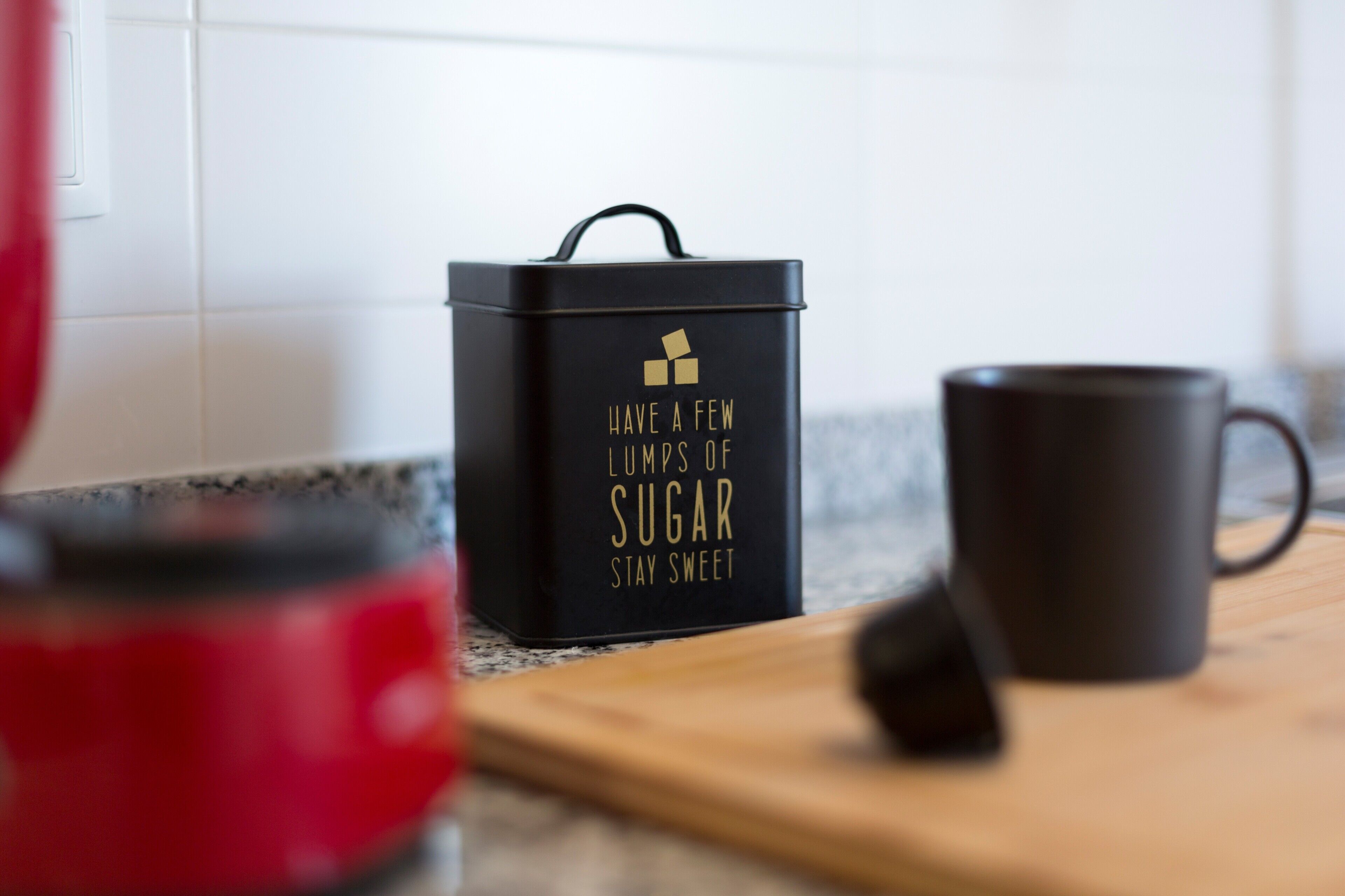 Uma bancada de cozinha com um recipiente preto, um eletrodoméstico vermelho e uma caneca preta.