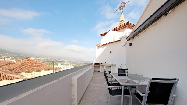 Un balcón con mesa y sillas, vistas de la ciudad y un edificio con un tejado peculiar.