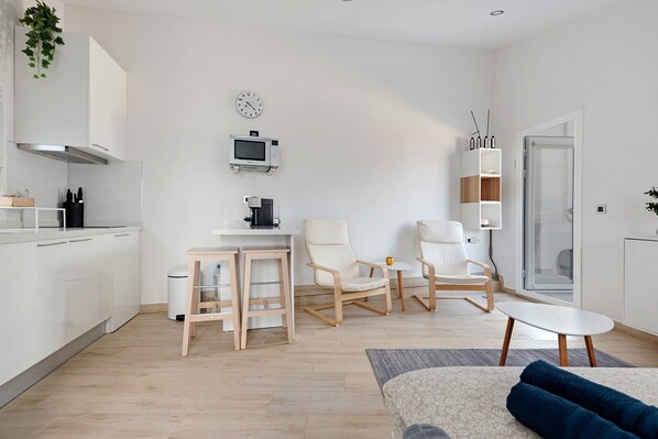 Une cuisine moderne avec des meubles blancs, un four à micro-ondes et une machine à café. On y trouve deux chaises blanches, une petite table et un canapé bleu.
