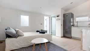 Habitación pequeña con cama, una mesita, espacio para la cocina y una puerta que da a otra habitación.