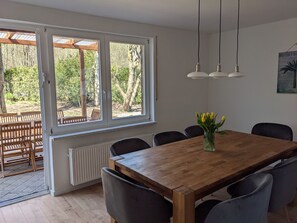 A dining area with a wooden table and chairs, large windows with a view of trees, and pendant lights. - House Icefen (Blankenheim)