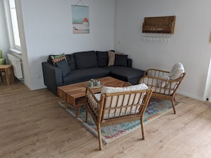 A cozy living room with a dark grey sectional sofa, a wooden coffee table, and a wicker chair. A colorful rug is on the floor, and a small table with a plant is visible in the corner.