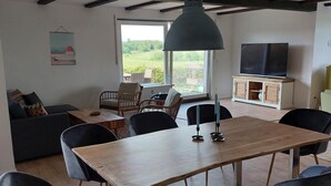 En matplats med ett träbord och svarta stolar, en tv på ett skåp och utsikt över grönområden utanför.
