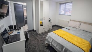A compact hotel room with a bed, a desk with a laptop, a wardrobe, and a window with blinds.