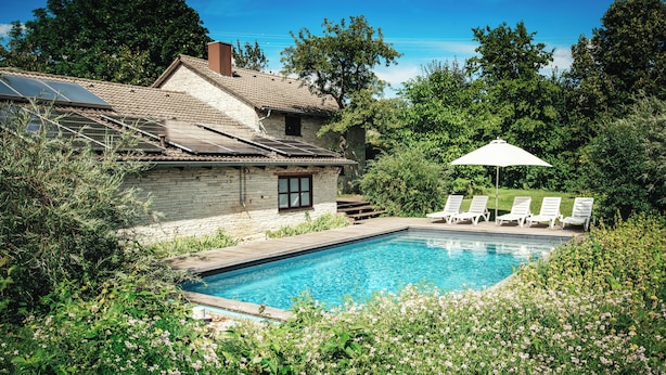 Una piscina con una casa de piedra, rodeada de vegetación y un paraguas blanco.