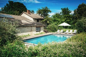 Een zwembad met een stenen huis, omringd door groen en een witte parasol.