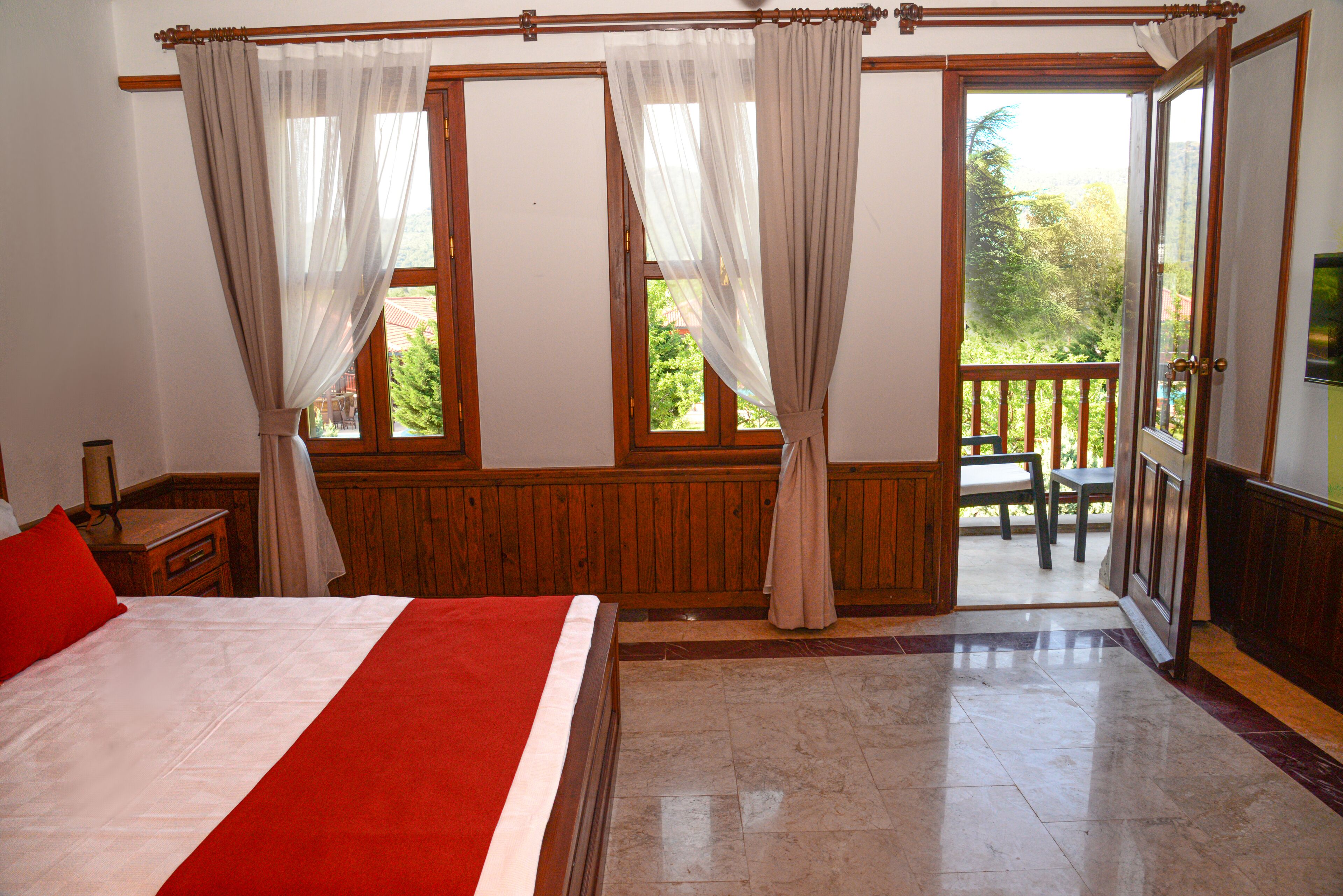 Ein Schlafzimmer mit einem großen Bett, Holzpaneelen, einem Balkon mit Tisch und Stühlen sowie leichten Vorhängen an den Fenstern.