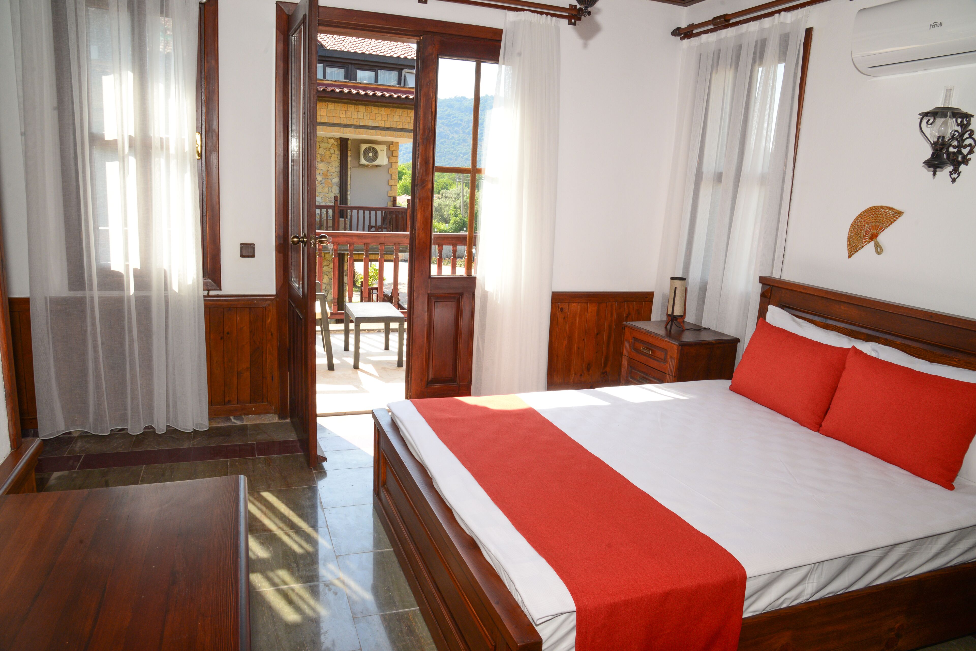 Een slaapkamer met een houten bed, een rood-wit beddeken, een nachtkastje van hout en uitzicht op een balkon.