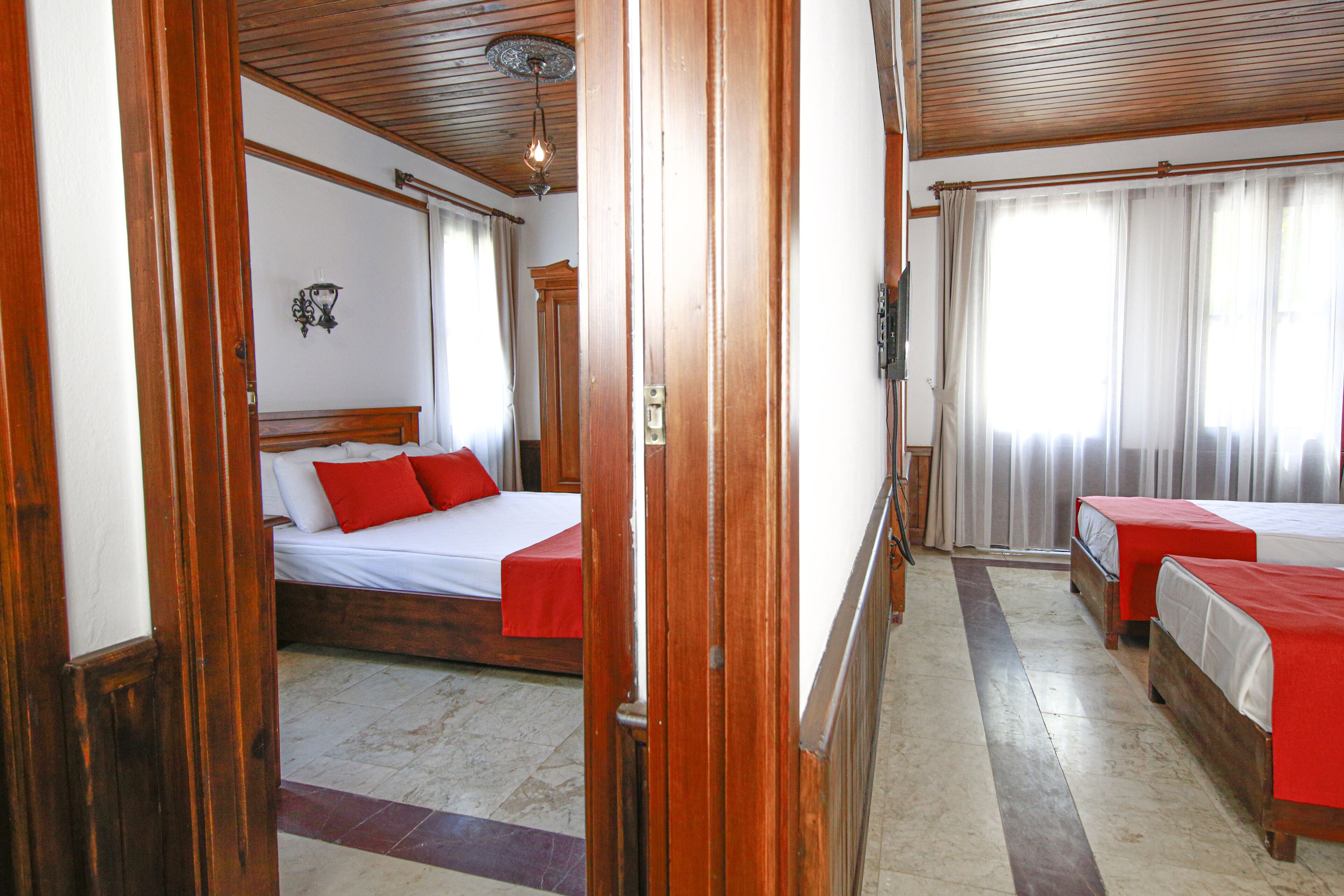 A hotel room with a wooden floor, a bed with red bedding, a bedside table, and a window with curtains.