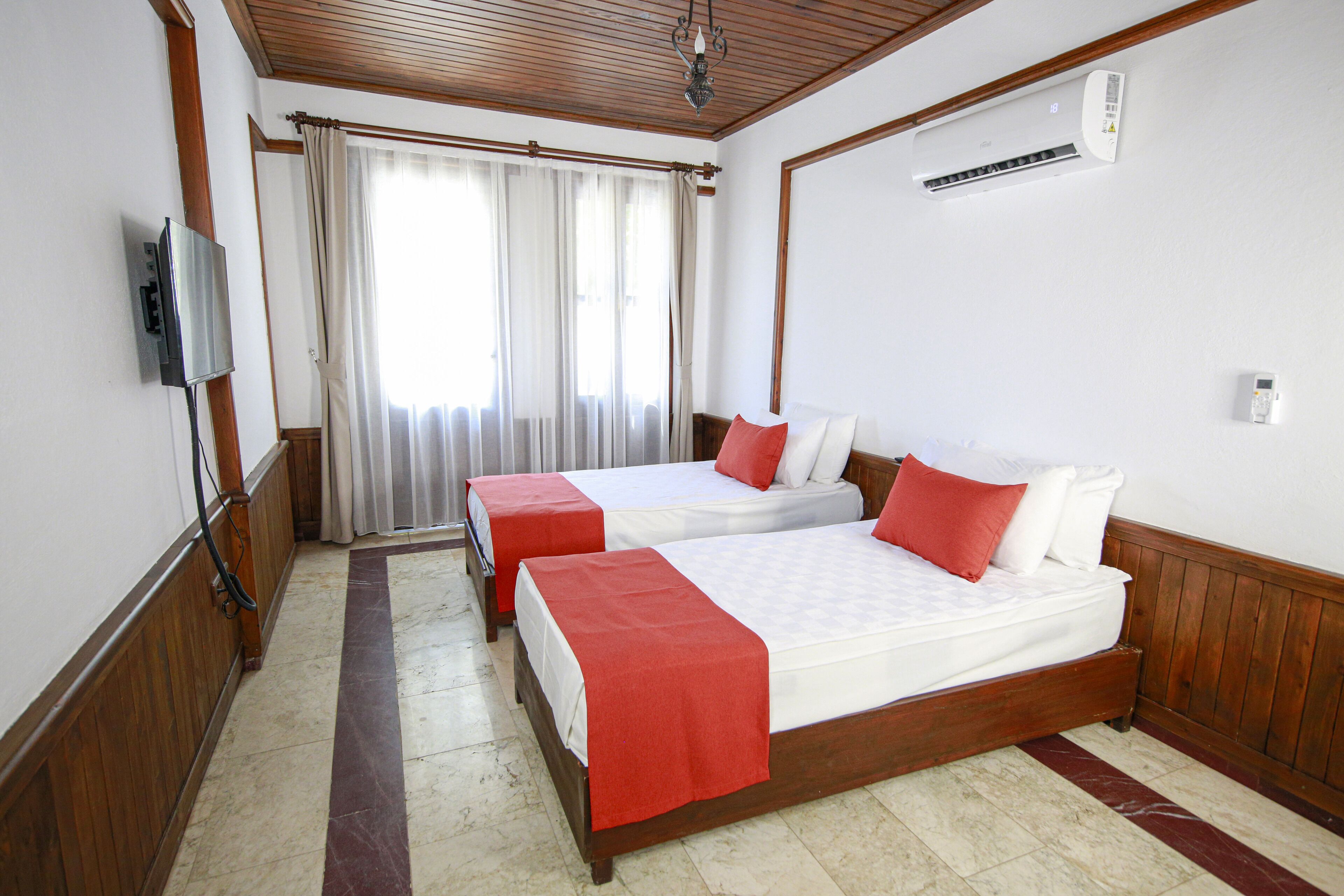 A hotel room with two beds, wooden paneling, a television, an air conditioning unit, and a window with curtains.