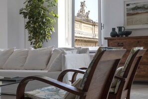 Una sala de estar con sofá, sillas, una planta, un ventanal con vista a una estatua y un mueble de madera con objetos decorativos.