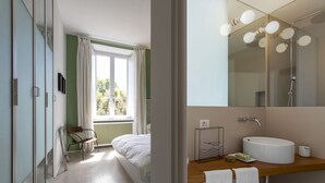 A modern bedroom with a bed, a chair, a window, and a bathroom with a sink and mirror. - GuestHost - Large and bright 200sqm apartment, able to accommodate up to 7 people, located on the 3rd floor with elevator (not suitable for disabled people) of a building dating back to the 19th century. The property overlooks Piazza Fanti's garden and i (Roma)