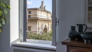 A room with a view of a historic building through a window, a wooden cabinet with kitchenware, and a plant on the left. - GuestHost - Large and bright 200sqm apartment, able to accommodate up to 7 people, located on the 3rd floor with elevator (not suitable for disabled people) of a building dating back to the 19th century. The property overlooks Piazza Fanti's garden and i (Roma)